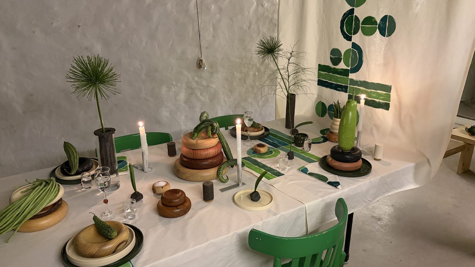 Mesa larga con mantel blanco, piezas de cerámica, cactus y textil verde colgado detrás
