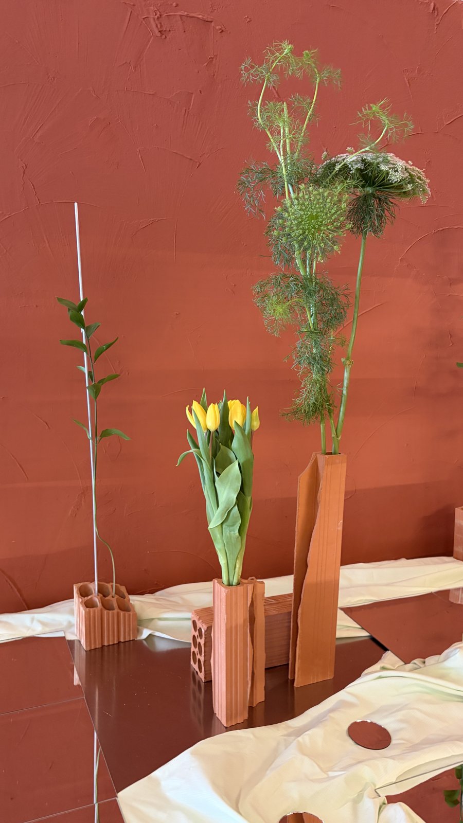 Floreros de ladrillo con tulipanes amarillos y ramas sobre espejos, contra una pared terracota